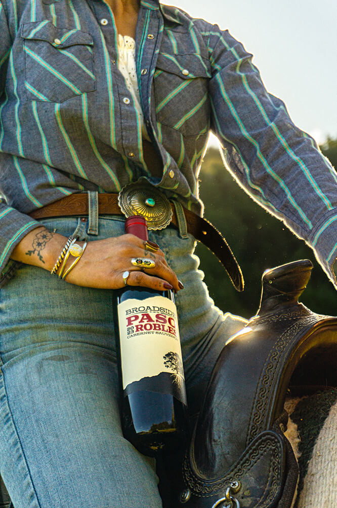 a lady riding a horse holds a bottle of 2023 cabernet sauvignon near her lap of the saddle