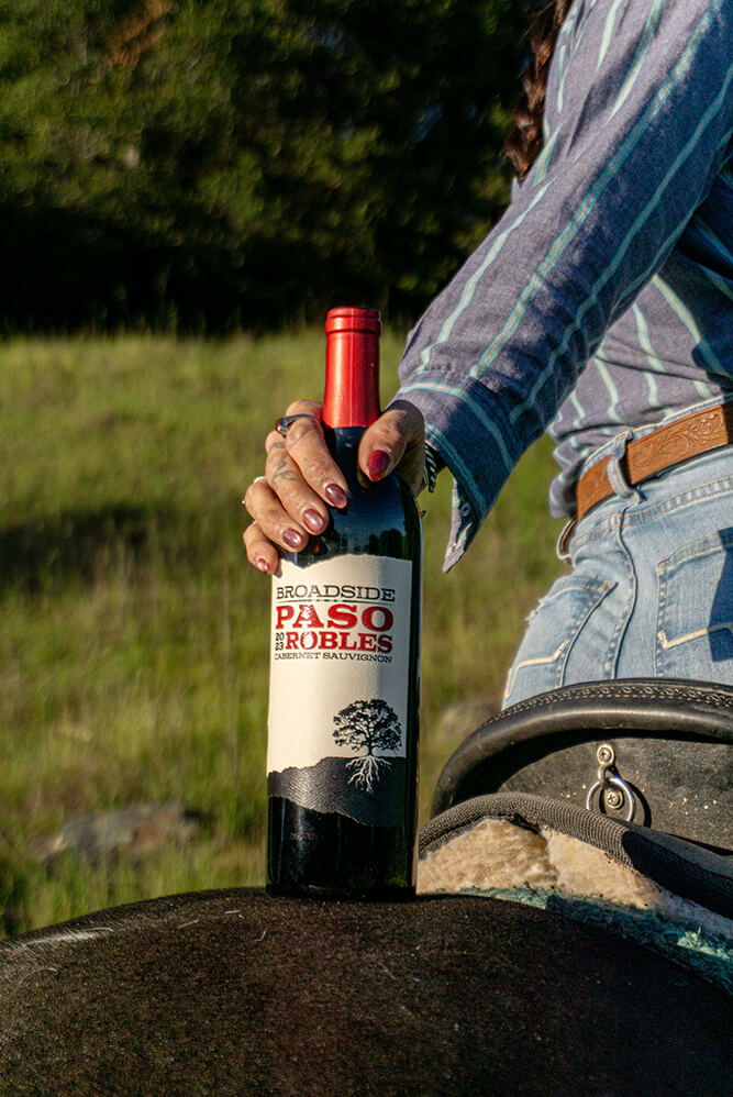 a lady holds a bottle of 2023 cabernet sauvignon upon the rear of a horse she is riding