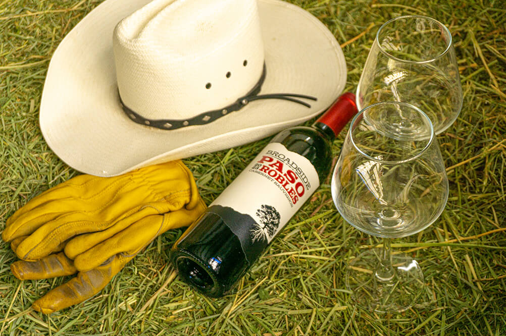 A bottle of 2023 Cabernet Sauvignon on hay next to two empty glasses, a white cowboy hat, and some yellow leather gloves.