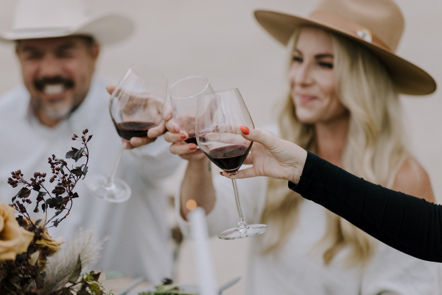a close-up of three individuals toasting wine glasses between a man and lady wearing cowboy hats and another lady out of view